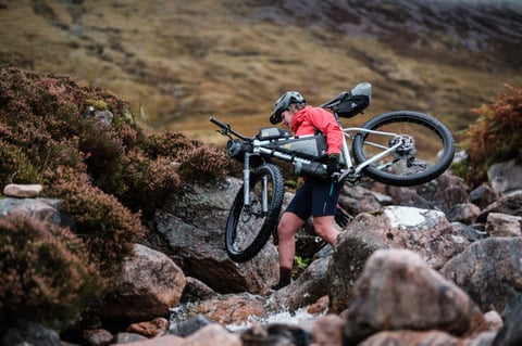 Rider carries a mountain bike loaded with Apidura Backcountry bags across a rocky stream below heather-covered hills.