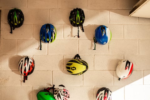 Assorted Scott cycling helmets hang on a white breeze-block wall lit by low sun.