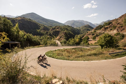 Two riders lean through a hairpin bend on a quiet mountain road with wooded Pyrenean peaks behind.