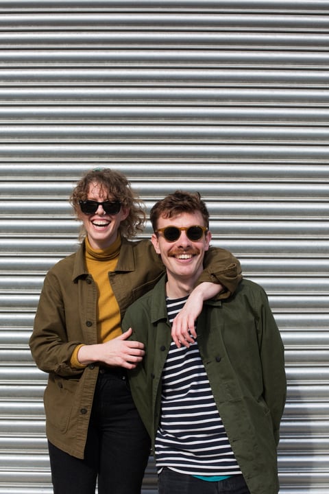 Wizard Works founders Harry and Veronica laughing in sunglasses in front of a metal roller shutter.