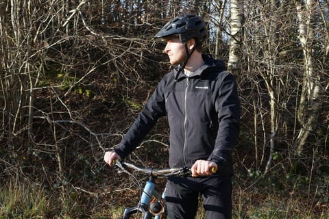 Rider in the black Endura MT500 Waterproof Jacket II holds the handlebars of a mountain bike in winter woodland.