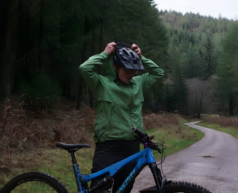Rider in a muddy green Alpkit Balance waterproof jacket pulls on a helmet beside a blue mountain bike on a forest track.