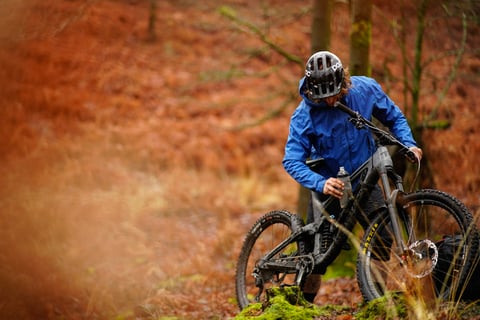 Rider in a blue dhb Trail waterproof jacket stands with a mountain bike in autumn woodland.