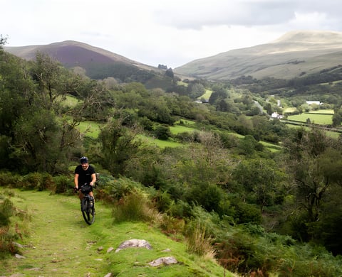 Bikepacker rides a grassy track above a green valley and hills in Wales, on one of the UK's bikepacking routes.
