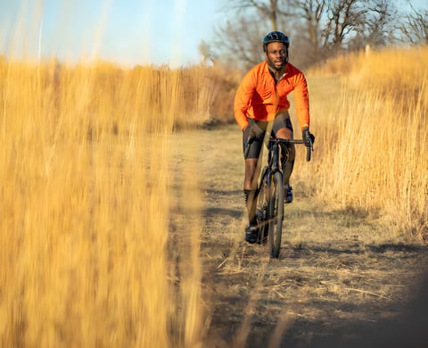 Rider in an orange jacket on a 2021 Salsa steel bike along a dirt trail between tall golden grasses.