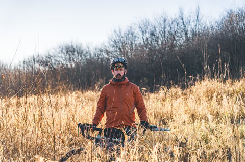 Rider in the orange 7mesh Copilot waterproof jacket standing with a gravel bike in tall dry grass.