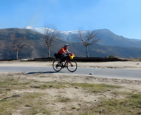 Rider on a loaded red bike on a quiet road below snow-capped mountains while bikepacking the length of Spain.