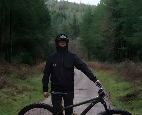 Rider in a black TSG Drop Rain Jacket and helmet stands with a mountain bike on a forest track.