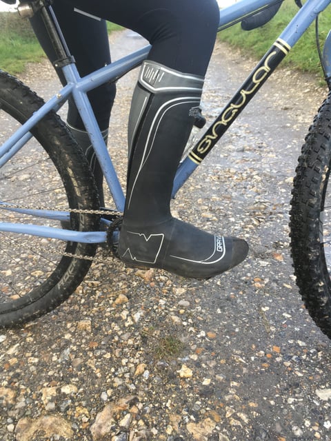 SPATZ GravlR overshoe covering a rider's lower leg, foot on the pedal of a blue bike on a wet gravel track.