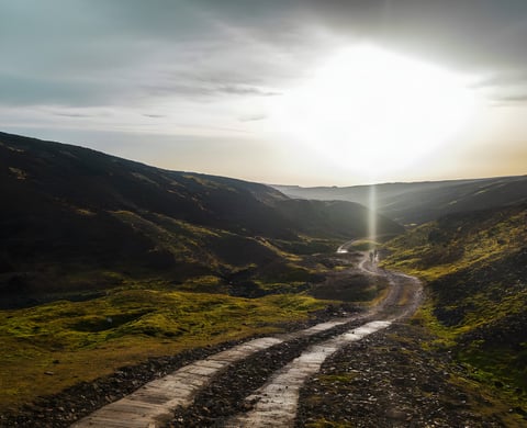 Gravel track winding through a moorland valley towards a low sun, with two distant riders, from Restrap's route catalogue.