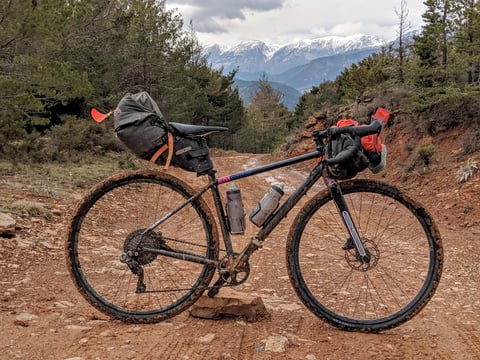 Mud-spattered gravel bike with Ortlieb seat pack and handlebar pack on a red dirt track below snowy mountains.