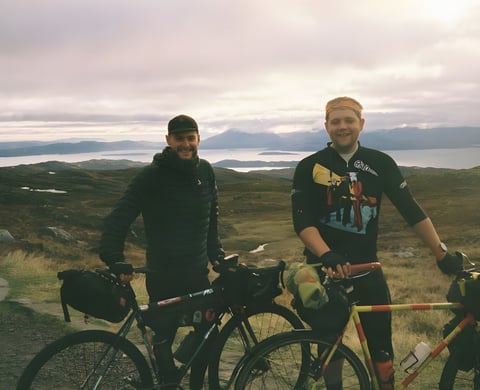 Two smiling riders with loaded touring bikes on a hilltop above a sea loch on a fixed-gear tour of north-west Scotland.