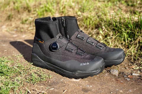 Fizik Terra Artica X2 winter boots with BOA dial and neoprene cuffs, standing on a dirt track beside grass.