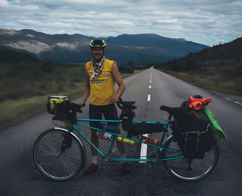 Rider stands beside a loaded teal tandem on an empty road in the Scottish Highlands on the NC500.