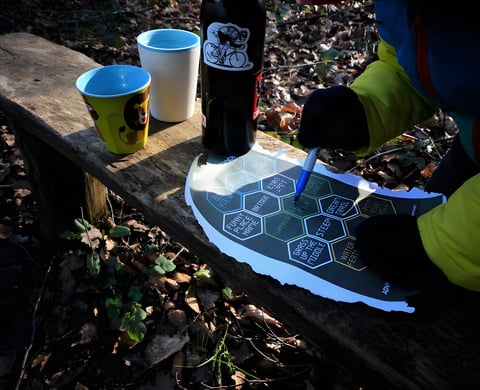 Rider ticks off The (Socially) Distance(d) 2021 hexagonal task card with a pen on a wooden bench beside mugs and a bottle.