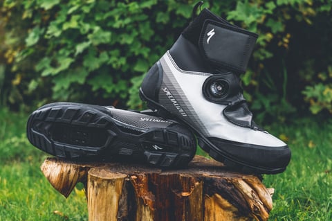 Specialized Defroster Trail MTB winter boots in reflective silver and black, one on its side showing the sole, on a log.