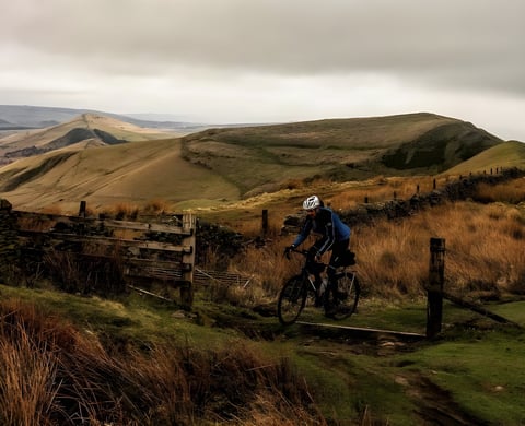 Rider on a gravel bike on a track near Macclesfield with Peak District hills and a wooden gate behind.