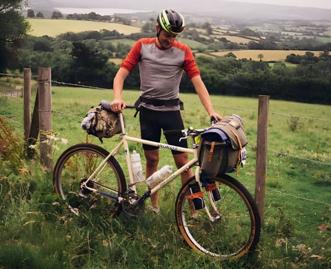 Rider with a vintage Specialized Rockhopper mountain bike loaded with bikepacking bags beside a fence in farmland.