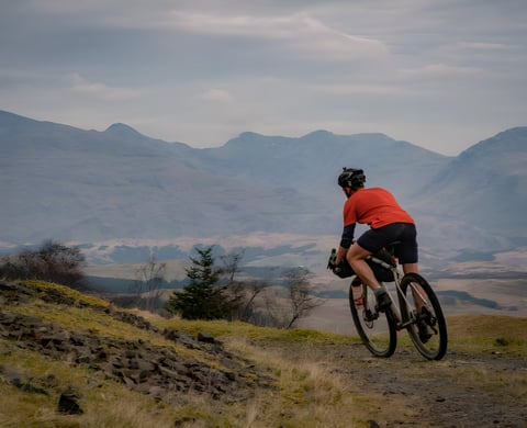 Rider in an orange top rides a rocky gravel track with Argyll hills behind, on a bikepacking overnighter.
