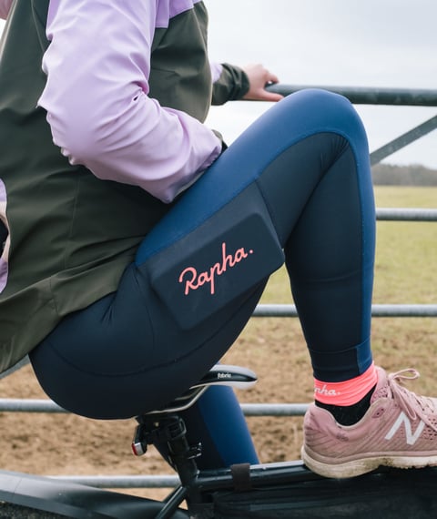 Rapha All Day Leggings in navy with a coral logo on the thigh pocket, worn by a rider sitting on a bike beside a gate.