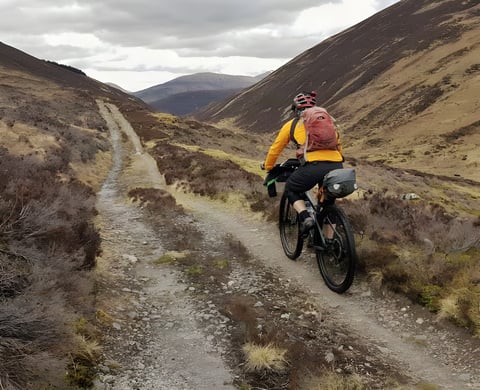 Rider with a backpack and loaded bike rides a rocky Highland track on the Central Highlander bikepacking route, Scotland.
