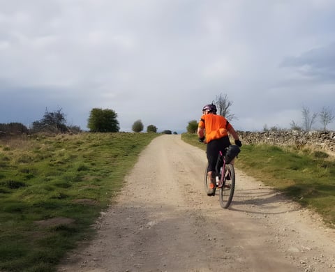 Dales Divide rider in an orange jersey climbs a gravel track between dry stone walls and rough grass.