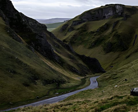Two cyclists ride a narrow road through a steep-sided rocky valley, promoting the 12HoursOfMay cycling challenge.