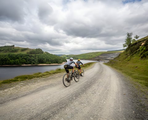 Two Gritfest riders on a wide gravel track beside a reservoir under cloudy skies in the Cambrian Mountains.