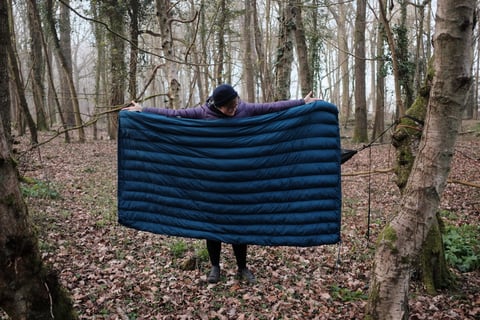 Camper holds up the blue baffled Alpkit Cloud Cover goose down quilt between trees, a hammock hanging beside it.