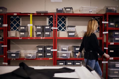 Restrap worker walks past red shelving stacked with labelled plastic boxes of cut bag parts.