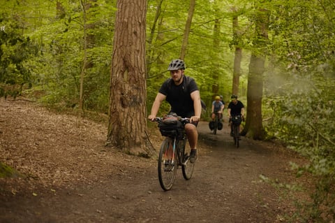 Rider with a handlebar bag leads two others along a muddy path through Leeds woodland.