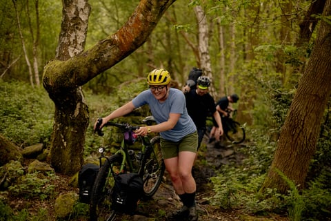 Riders push gravel bikes fitted with Restrap panniers up a rocky woodland trail.