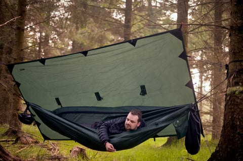 Camper lies in a green hammock beneath a tarp pitched among conifers at the overnight camp near Swinsty.