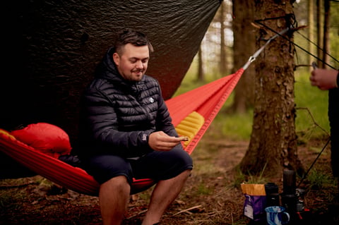 Camper in a puffer jacket sits in a red hammock under a tarp, eating a snack at the Swinsty campsite.