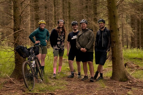 Five Restrap overnighter riders pose in pine forest, one with a loaded gravel bike propped against a tree.