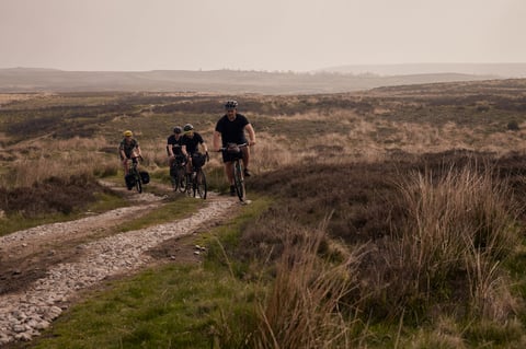 Four riders on loaded gravel bikes climb a stony double track across hazy open Yorkshire moorland.