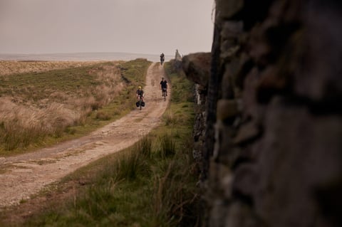 Riders follow a rough moorland track beside a dry stone wall above Ilkley, one with rear panniers.