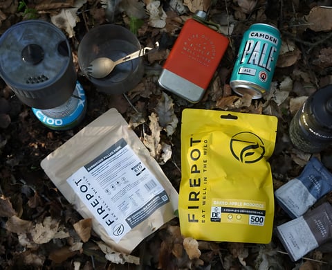 Firepot dehydrated meal packets laid out in leaf litter with a camping stove, spork, flask and a can of beer.