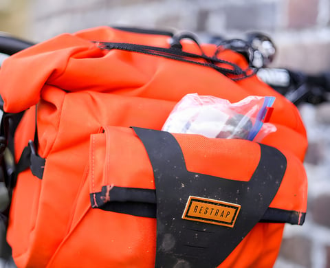 An orange Restrap Bar Pack mounted on handlebars against a brick wall.
