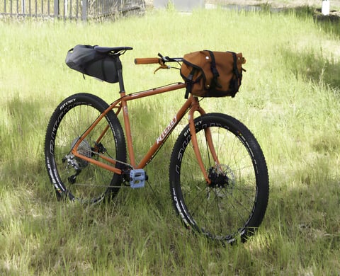 A loaded Ritchey Ascent bike with a saddle bag and handlebar bag parked in long grass.