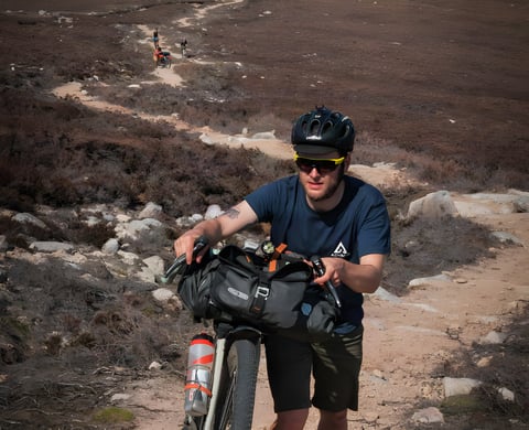 Rider in Vulpine gravel shorts pushes a bikepacking bike with a handlebar bag up a rocky heather track in Scotland.