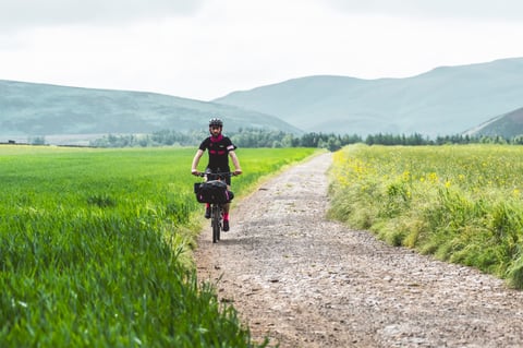 Rider on a loaded bike on a stony farm track between green fields and hills on the Rapha Pennine Rally.