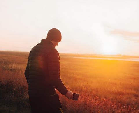Backlit camper in a beanie and down jacket holds a mug, watching low sun over open marsh on a midweek local overnighter.