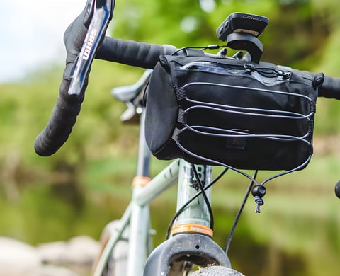 Close-up of the Ashdown Bags bar bag strapped under a gravel bike's handlebars, elastic cord on the front, beside a lake.