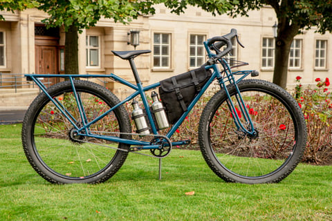 Clandestine Long Tail in blue steel with a long rear rack, drop bars, a frame bag and two steel bottles on grass.