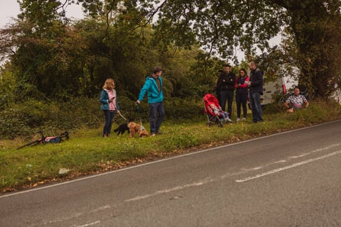 Spectators with two dogs and a pushchair wait on a grass verge for the Women's Tour of Britain, a bike laid on the grass.