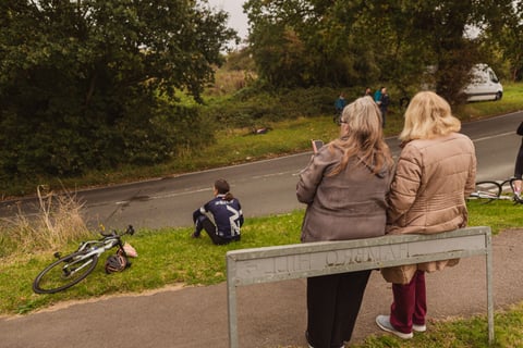 Two women lean on the Hambro Hill road sign watching the Women's Tour of Britain route, bikes left on the grass.