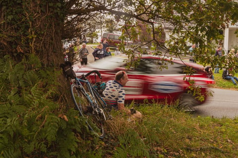 Spectator in a spotted club jersey sits under a tree as a red Women's Tour of Britain convoy car blurs past.