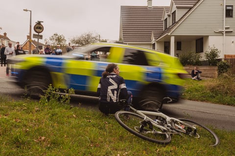 A police car blurs past a spectator sat on a grass verge beside a bike at the Women's Tour of Britain in Essex.
