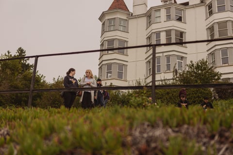 Two women chat on a grassy bank below a tall white building as spectators wait for the Women's Tour of Britain.
